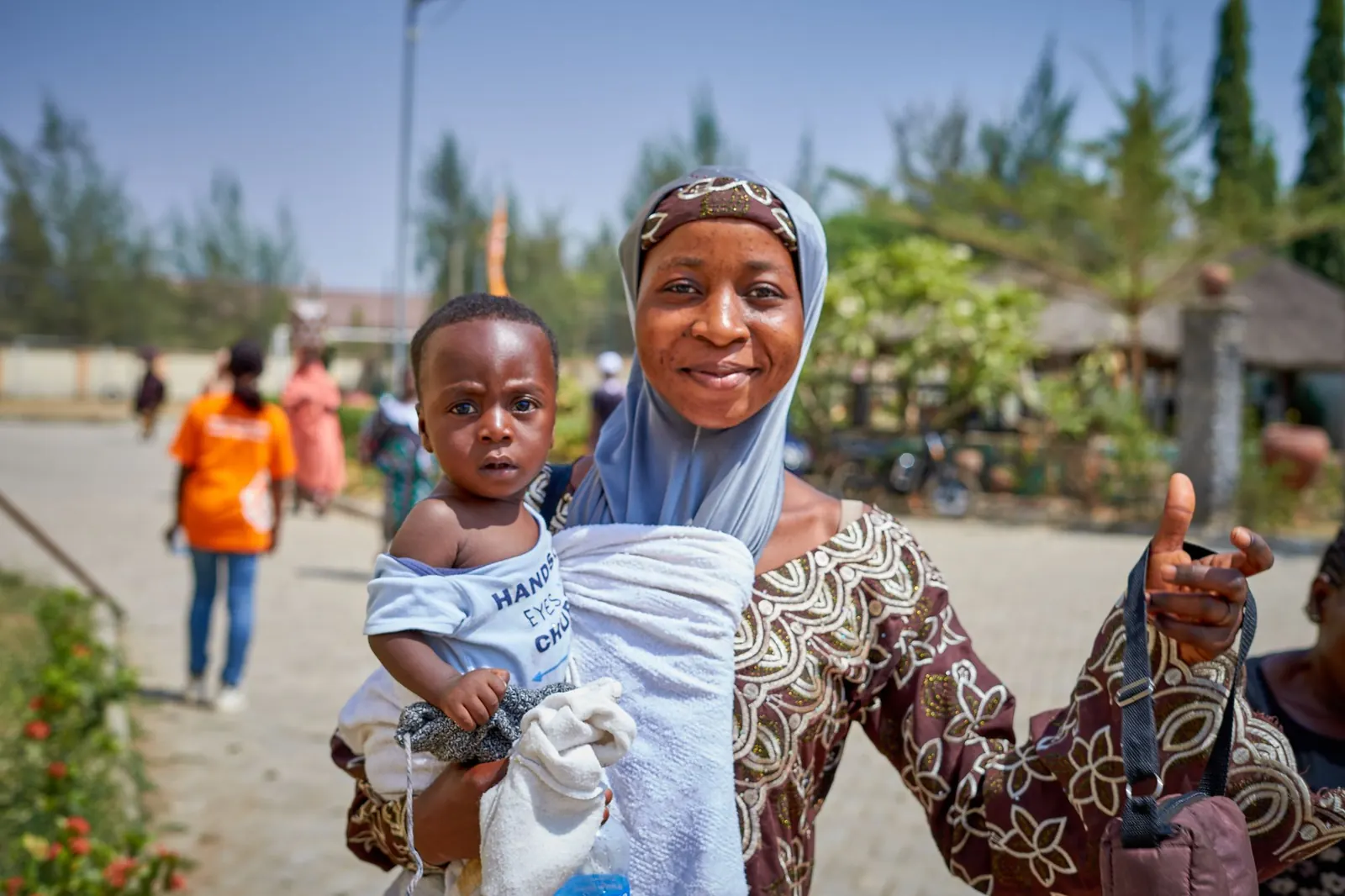 Smiling Nigerian mother in a headscarf holding her baby boy outside a clinic, giving a thumbs-up